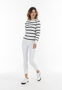 Fehér csíkos pulóver fekete csíkokkal, fehér, slim-fit farmernadrággal és fekete-fehér sportcipővel párosítva. A modell egy sima háttér előtt áll.
