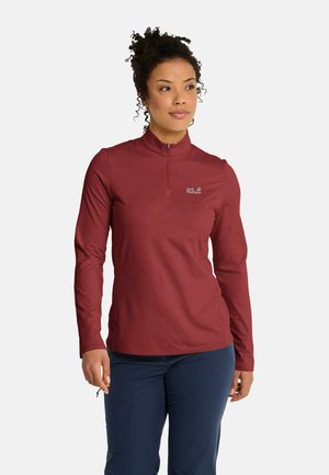 Donna con capelli ricci che indossa una maglia bordeaux con zip e maniche lunghe e pantaloni blu scuro, in piedi e che guarda di lato.