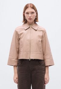 Trumpas smėlio spalvos odinis švarkas su smaigaluota apykakle, priekiu su užtrauktuku ir briaunotais akcentais apačioje bei rankogaliuose. Modelis dėvi tamsiai rudus kelnes.