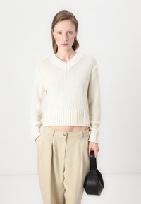 Kremfarget v-hals genser med teksturert strikk, ribbestrikket kant og lange ermer, kombinert med høy midje beige bukser og en svart håndveske.