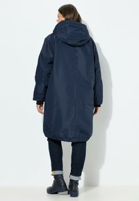 Parka impermeable de color azul marino con capucha, mangas largas, dobladillo elástico y costuras visibles, combinada con vaqueros oscuros y botas negras hasta el tobillo.