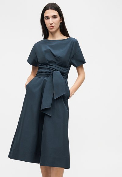 Γυναίκα με μακρύ μπλε φόρεμα με κοντά μανίκια, φαρδιά δεμένη ζώνη και τσέπες, στέκεται μπροστά σε απλό φωτεινό φόντο.