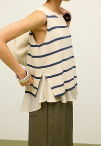 Beżowa bluzka bez rękawów w granatowe paski, z wysokim tyłem i rozcięciami z boku. W połączeniu z oliwkowo-zielonymi spodniami o szerokich nogawkach. Teksturowana torba.