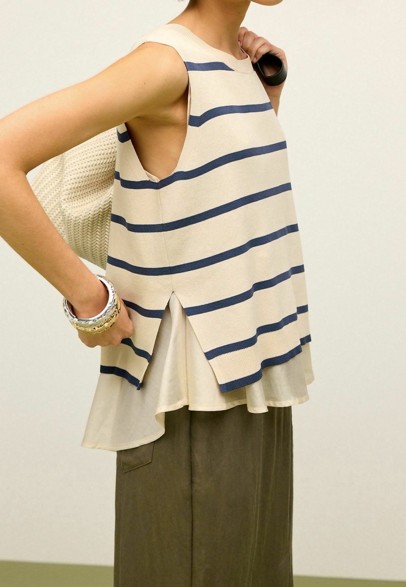 Beżowa bluzka bez rękawów w granatowe paski, z wysokim tyłem i rozcięciami z boku. W połączeniu z oliwkowo-zielonymi spodniami o szerokich nogawkach. Teksturowana torba.
