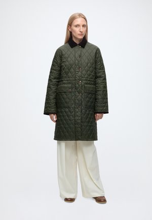 Woman wearing a long, quilted dark green coat with black collar, white wide-leg pants, and brown shoes, standing against a plain white background.