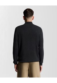 Mörkgrå stickad tröja med hög krage, långa ärmar och ribbade manschetter, visad på en person som bär beige byxor.