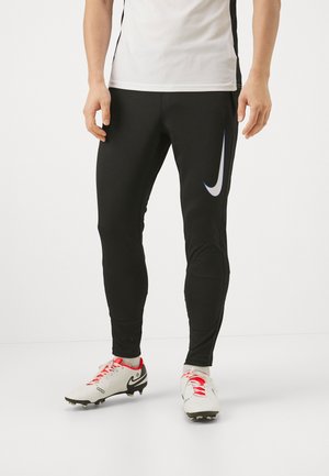 Persoon draagt zwarte sportbroek met een witte Nike-swoosh en witte voetbalschoenen met rode veters, staand op een witte vloer.