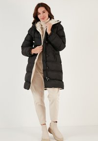 Black puffer coat with a cream interior, front zipper and snap closures, featuring a hood and elastic cuffs. Worn over beige pants and boots.