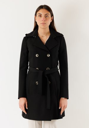 Donna che indossa un cappotto doppiopetto nero con bottoni dorati e cintura in vita, in piedi davanti a uno sfondo bianco semplice.