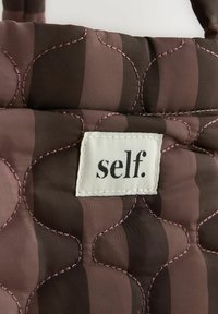 Quilted Tasche in Brauntönen mit einem wellenförmigen Muster und rosa Stichen. Verfügt über ein weißes Stoffetikett mit "self." in Schwarz gedruckt.