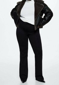 Persoon in zwarte high-waisted broek, zwart leren jack met bontkraag, witte geribbelde top en zwarte puntige schoenen, staande met handen op de heupen.