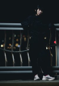 Femme en vêtements sombres appuyée sur une rambarde métallique la nuit, avec les lumières floues de la ville en arrière-plan, portant des baskets blanches avec des détails roses.