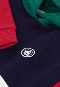 Marineblå genser med røde og grønne detaljer; har et sirkulært brodert logo. Materialet virker mykt med en teksturert overflate.