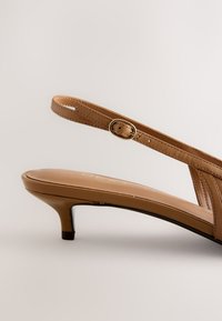 Sandale à bride arrière en cuir marron avec petit talon kitten et bride réglable avec boucle dorée sur fond clair.