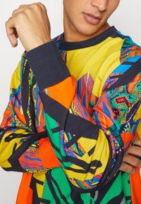 Chemise à manches longues colorée présentant des motifs vibrants en jaune, orange, vert et bleu, avec des dessins texturés et un col côtelé bleu marine.