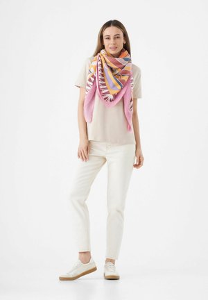 Kleurvolle sjaal met geometrische patronen in roze, oranje en paars, gedragen over een beige shirt, gecombineerd met witte jeans en lichte sneakers.