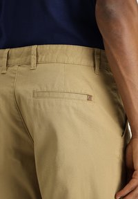 Pantaloni khaki con una texture liscia, dotati di una tasca posteriore e di un piccolo etichetta del marchio. I passanti per la cintura sono visibili in vita.