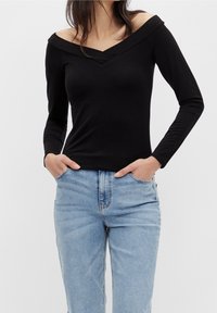 Frau in schwarzem, schulterfreiem Langarmoberteil mit hellblauen, hochgeschnittenen Jeans, Hände in den vorderen Taschen, vor einem schlichten Hintergrund.