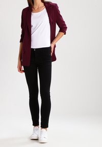Bordeaux blazer, witte t-shirt, zwarte slim-fit jeans en witte sneakers. Blazer heeft een reverskraag en opgerolde mouwen, wat een stijlvolle uitstraling creëert.