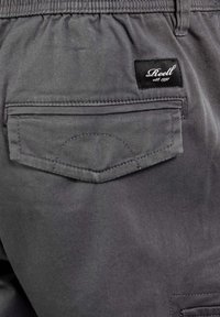 Graue Stoffshorts mit einer Gesäßtasche mit Klappe und einem schwarzen Etikett mit weißem Text. Glatte Textur und elastischer Bund.