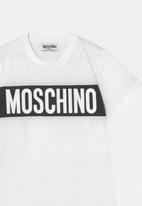 Bílý tričko s černým horizontálním pruhem a tučnými bílými nápisy "MOSCHINO". Vyrobeno z měkké bavlny, s kulatým výstřihem a krátkými rukávy.