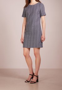 Šedé šaty s krátkým rukávem a texturovaným kostkovaným vzorem, střižené na tělo, ke kolenům, v kombinaci s černými kotníkovými sandály na hladkém povrchu.