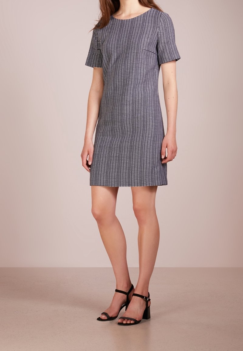 Šedé šaty s krátkým rukávem a texturovaným kostkovaným vzorem, střižené na tělo, ke kolenům, v kombinaci s černými kotníkovými sandály na hladkém povrchu.