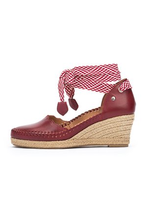 Zapato de cuña de cuero rojo con tacón de yute trenzado. Presenta una correa de tobillo tejida en rojo y blanco y costuras decorativas a lo largo de los bordes.