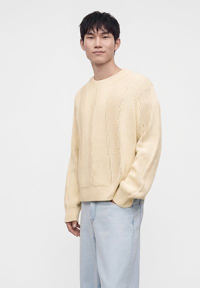 Jeune homme aux cheveux courts noirs portant un pull en maille crème et un jean bleu clair, debout avec une main dans la poche devant un fond uni.
