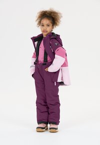 Lila und rosa isolierte Winterjacke mit passender Latzhose, ausgestattet mit verstellbaren Trägern, Taschen und warmer Fütterung. Braune Stiefel mit schwarzen Akzenten.