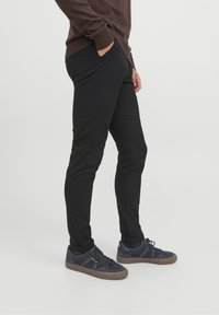 Personne portant un pantalon noir slim, des baskets bleu foncé avec des semelles marron, et un haut marron à manches longues, une main dans la poche, debout de profil.