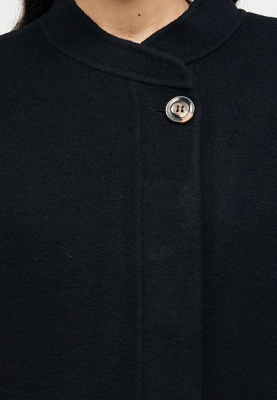 Μαύρο παλτό από μαλλί με ψηλό γιακά, που διαθέτει κλείσιμο με ένα μόνο κουμπί και υφασμάτινη υφή. Ξεχωρίζει για τις καθαρές γραμμές και τον μινιμαλιστικό σχεδιασμό του.