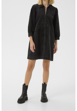 Frau trägt ein schwarzes Cord-A-Linien-Kleid mit 3/4-Ärmeln und einem Frontreißverschluss, kombiniert mit schwarzen klobigen Stiefeletten.