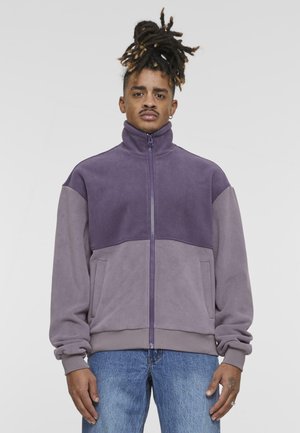 Purple zip-up fleece jacket featuring color-block design, soft texture, high collar, and front pockets, paired with blue denim jeans.