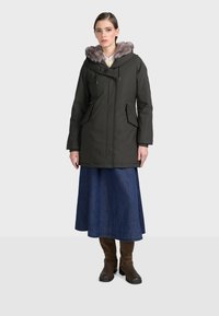 Ciemnozielona parka z kapturem wykończonym futrem, z dwoma bocznymi kieszeniami, na zamek, noszona na długą spódnicę dżinsową i brązowe botki.