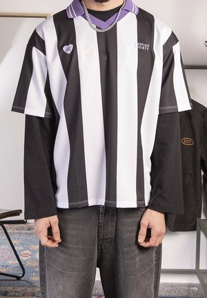 Persoon draagt een zwart-wit gestreept shirt met korte mouwen over een zwarte trui met lange mouwen, grijze spijkerbroek en een zilveren ketting.