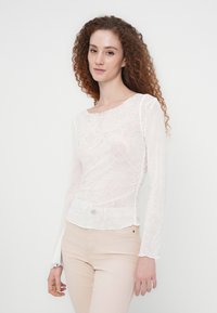 Νεαρή γυναίκα με μακριά σγουρά μαλλιά, που φοράει ένα διάφανο λευκό μακρυμάνικο τοπ με σχέδια και ανοιχτό μπεζ παντελόνι, στέκεται μπροστά σε ένα απλό φόντο.