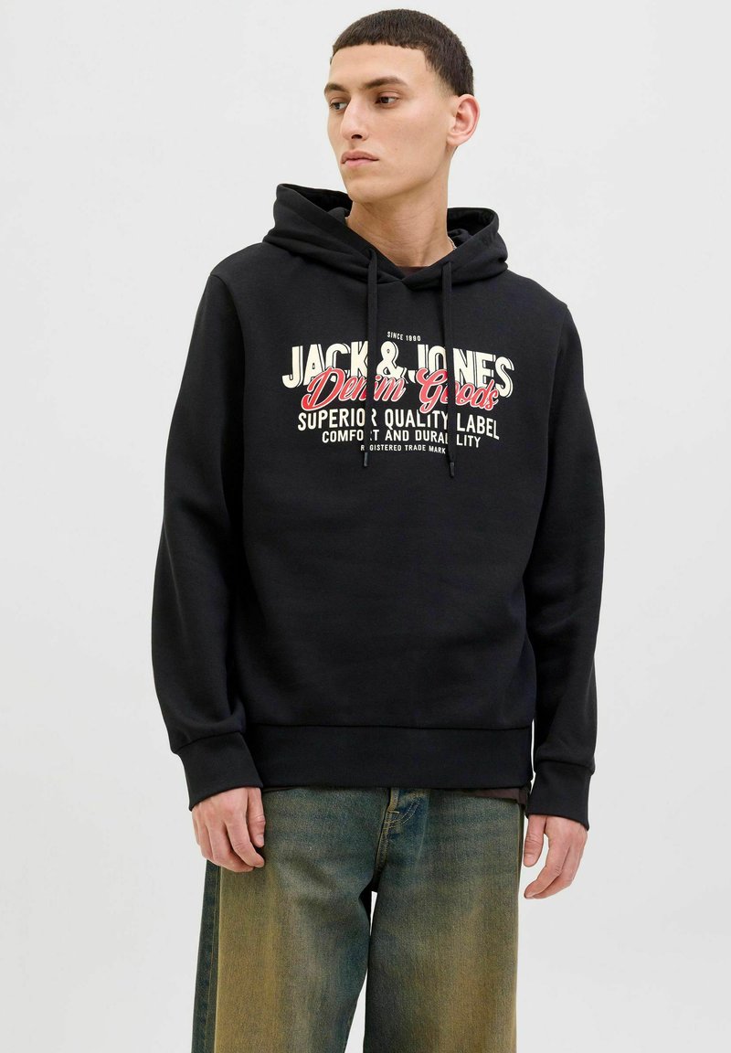 Sudadera negra con cordón, que presenta un gráfico frontal en texto blanco y rojo. Puños y dobladillo acanalados, combinada con denim claro.