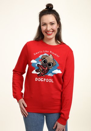Uśmiechnięta kobieta o ciemnych włosach związanych w kok, w czerwonej bluzie z nadrukiem kreskówkowego psa w kostiumie oraz napisem "Smells Like Trouble! DOGPOOL."