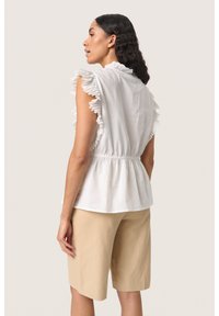 Hvid bomuldsbluse med flæser på ærmerne og elastisk talje, parret med beige shorts. Simpelt design med blød tekstur og diskret detaljer.