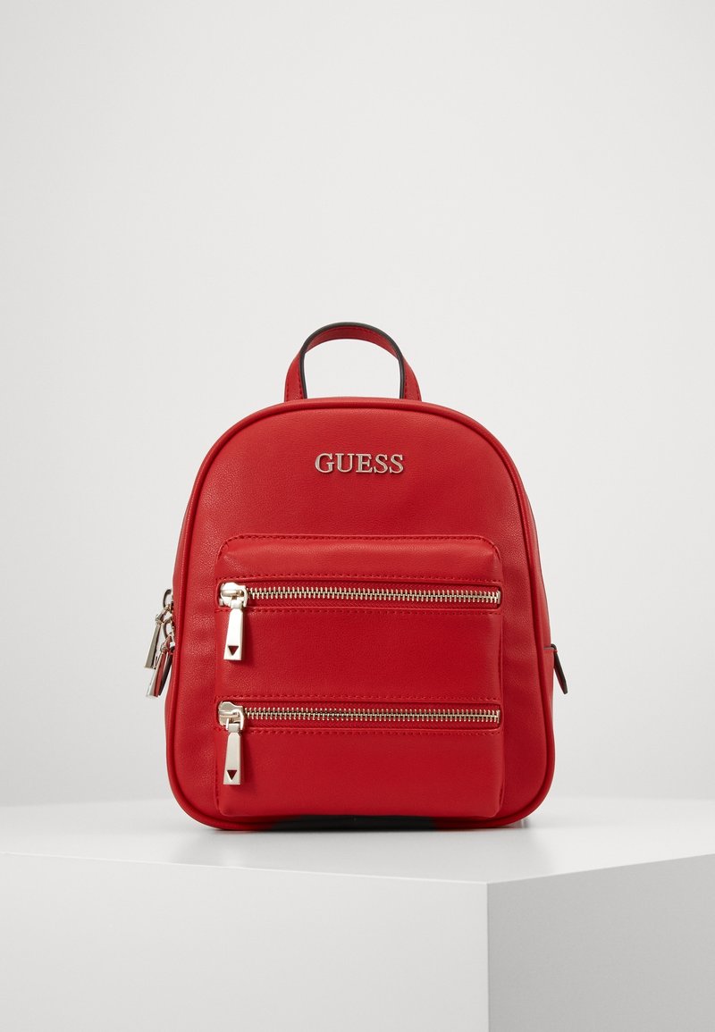 Sac à dos compact rouge Guess avec deux poches avant zippées et une poignée supérieure, présenté sur une surface blanche devant un fond uni.