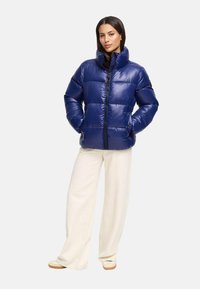 Niebieska kurtka puchowa z wysokim kołnierzem i zamkiem z przodu, z pikowaną konstrukcją. Model nosi kremowe spodnie o szerokich nogawkach i buty.