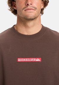 Bruine sweatshirt van katoen met een geribbelde ronde hals. Bevat een rood rechthoekig label met witte tekst "QUIKSILVER" en een logo.