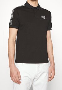 Svart pikétröja i bomull, med klassisk krage och tre knappar. Vita varumärkesdetaljer på ärmarna, mjuk textur.