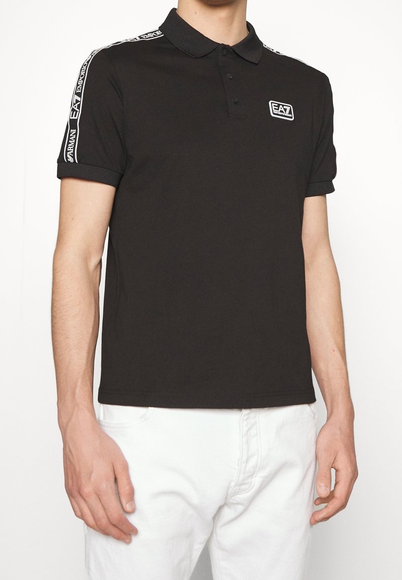 Svart pikétröja i bomull, med klassisk krage och tre knappar. Vita varumärkesdetaljer på ärmarna, mjuk textur.