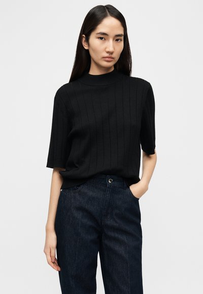 Μαύρο ριμπ πλεκτό πουλόβερ με κοντά μανίκια και ψηλό γιακά, συνδυασμένο με σκούρο τζιν παντελόνι, που διαθέτει κλασικές τσέπες και κούμπωμα με κουμπιά.