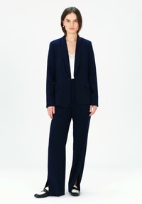 Marineblauer Maßanzug mit Blazer und weiten Hosen, die vorne Schlitze haben. Über einem weißen Oberteil mit Spitzenbesatz getragen. Schwarz-weiße Schuhe.