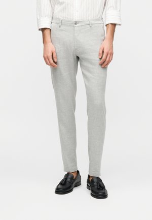 Mężczyzna noszący jasnoszare wąskie spodnie, białą koszulę w paski wpuszczoną w spodnie oraz czarne mokasyny z chwostami, stojący na białym tle.