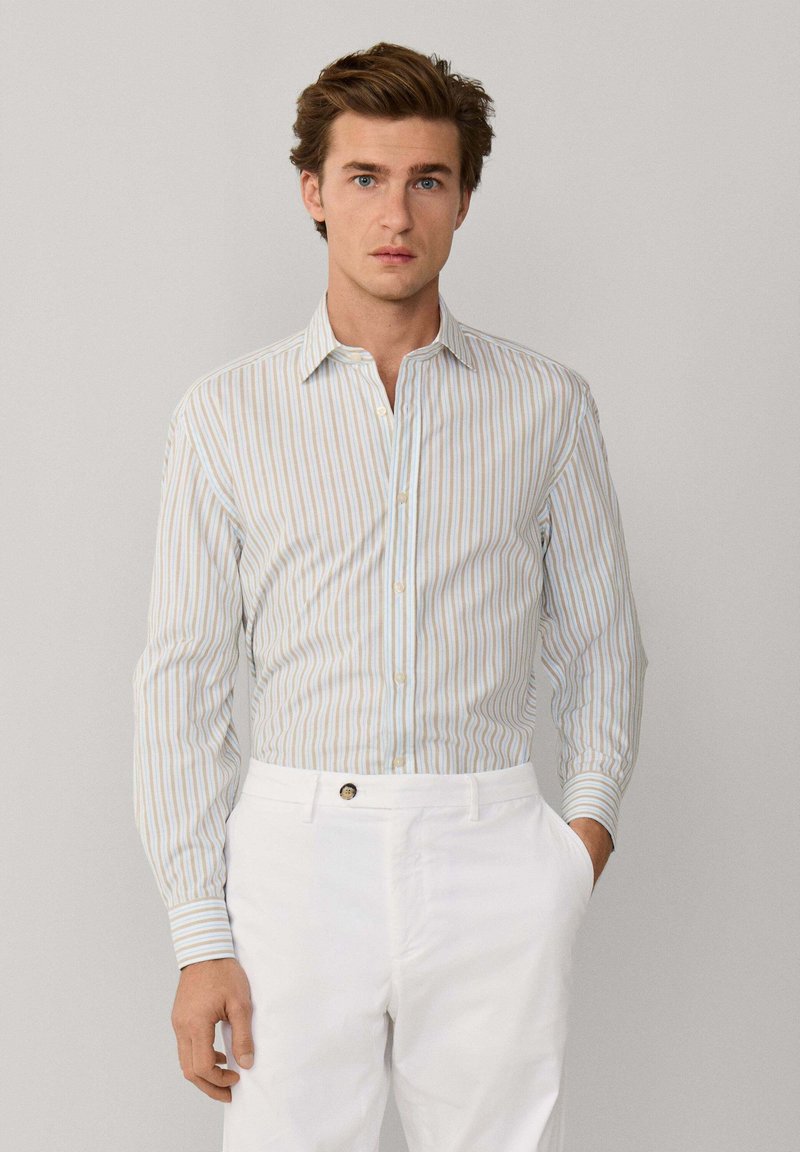 Mann mit hellbraunem Haar, der ein beiges, blau-weiß gestreiftes Hemd trägt, das in weiße Hosen gesteckt ist, mit einer Hand in der Tasche.