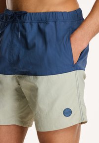 Man in twee-kleurige zwembroek met een marineblauwe bovenhelft en een lichtbeige onderhelft, hand in zak, logo-patch op het linkerbeen.